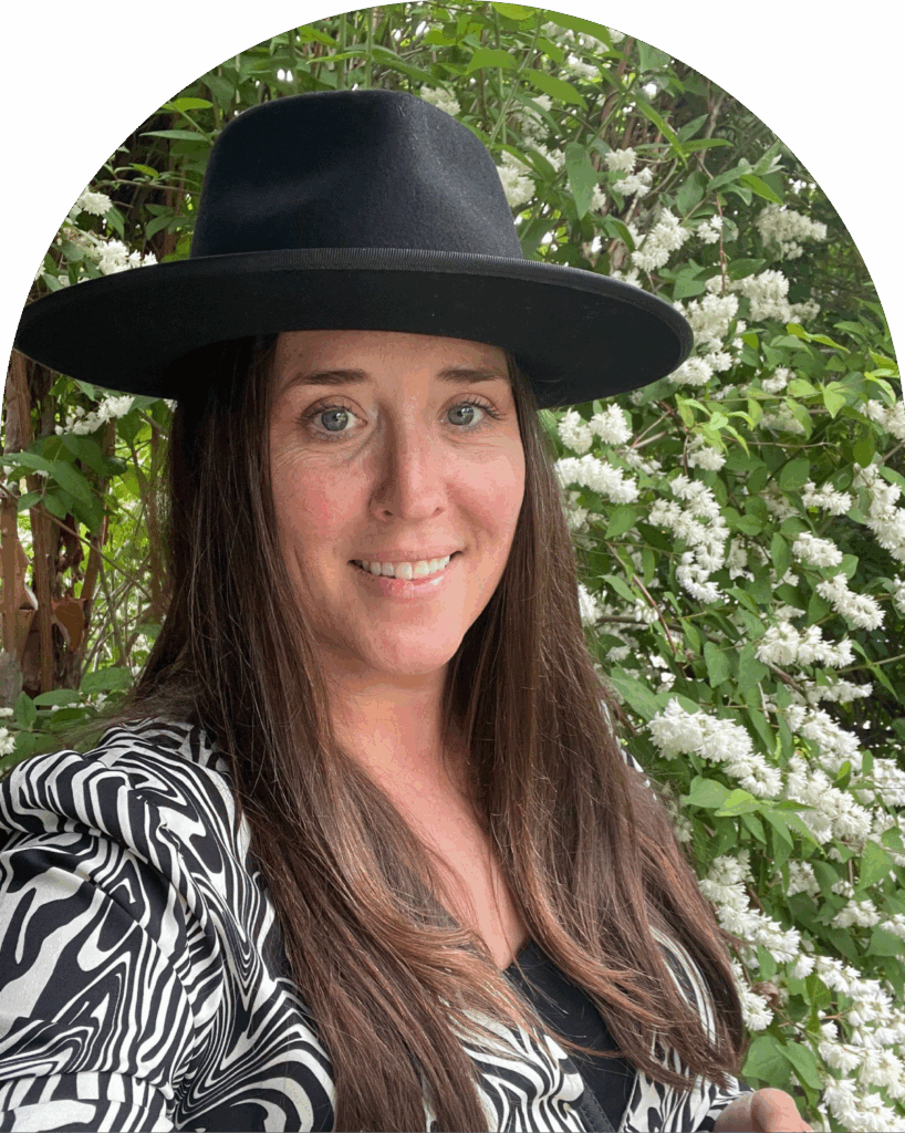Amber Sawaya smiling outdoors in front of a flowering hedge, wearing a wide-brim black hat and a black-and-white patterned blazer.
