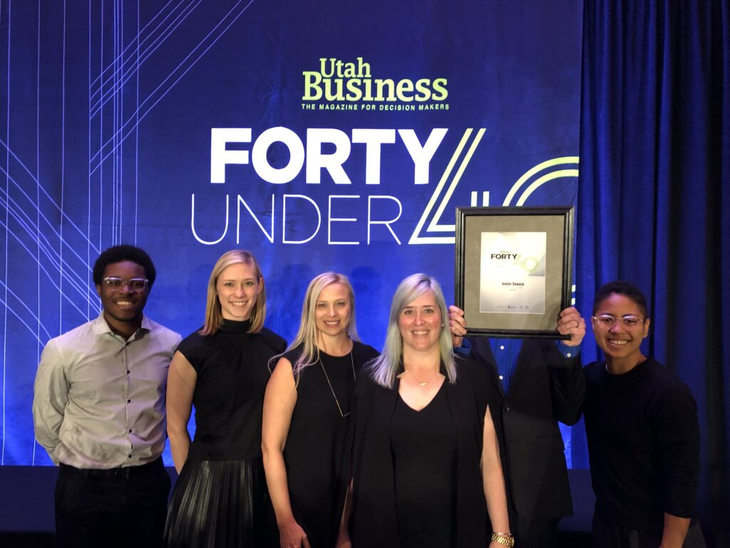 Amber Sawaya (center) with colleagues on stage at the Utah Business Forty Under 40 awards, smiling in front of the event backdrop as someone holds the framed award.